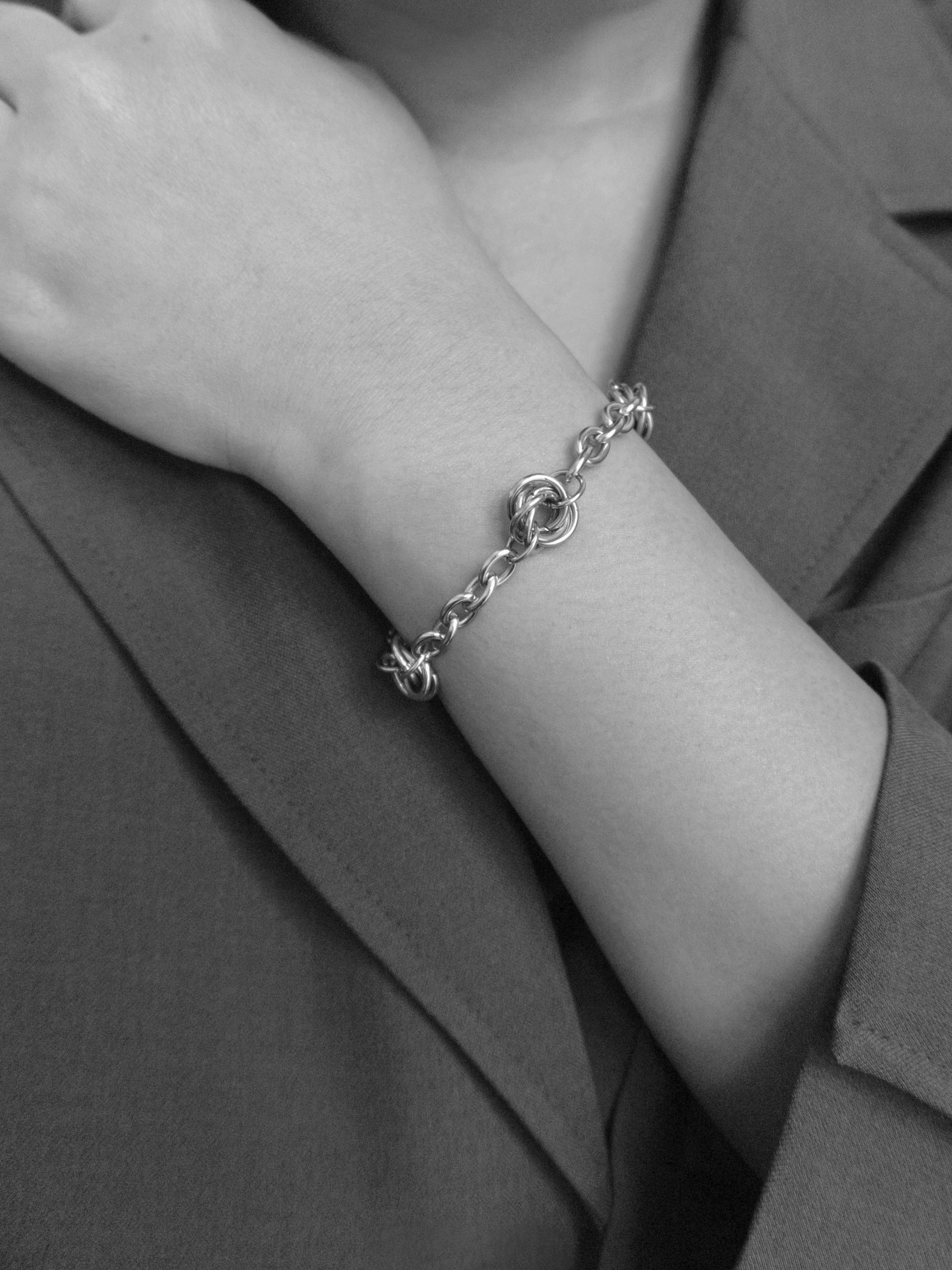 A close-up black and white photo of a delicate chain bracelet worn on a person's wrist, with arms crossed in the background.