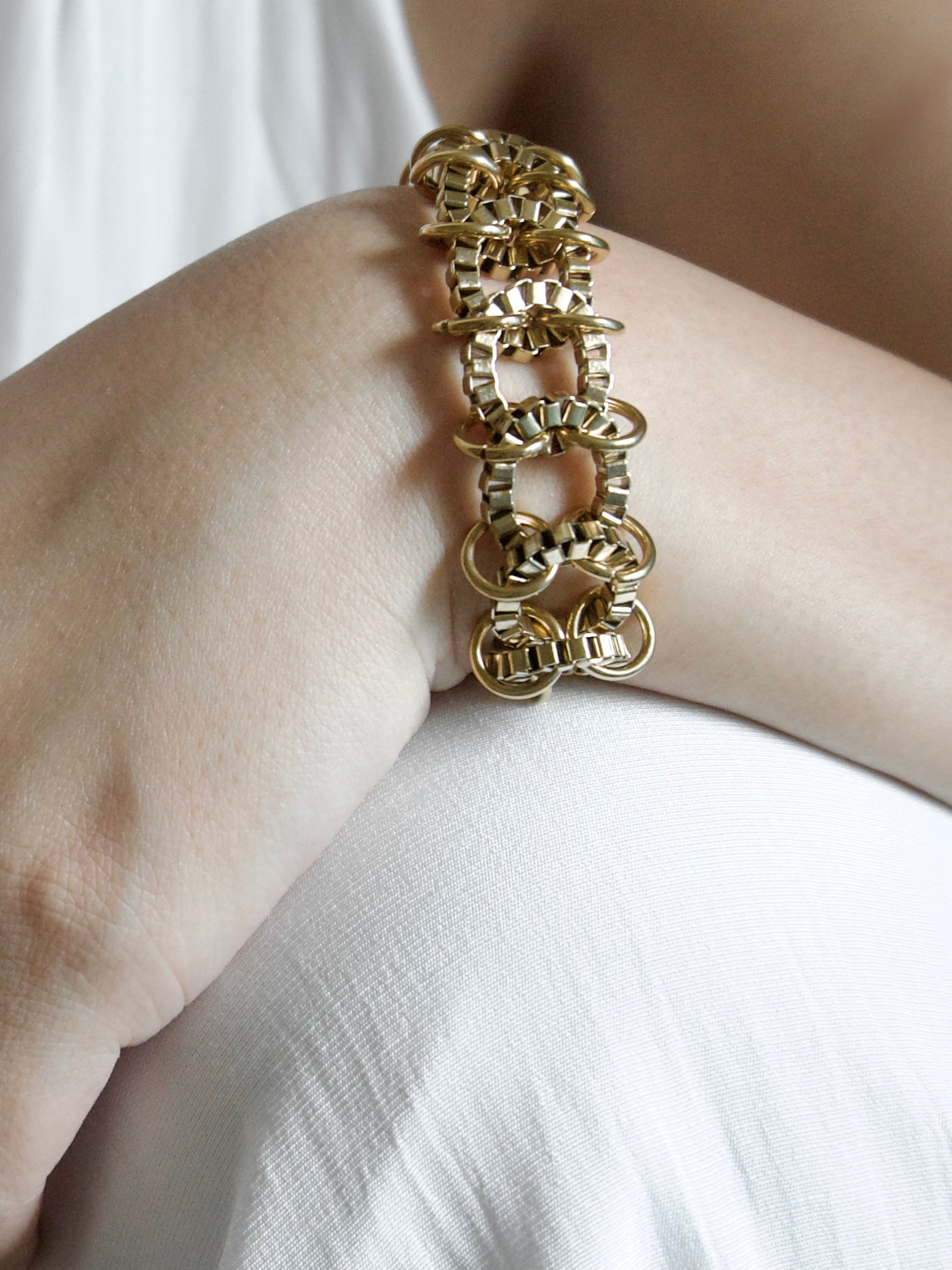 A close-up of a gold-toned chain-link bracelet worn on a person's wrist, with a white top visible in the background.
