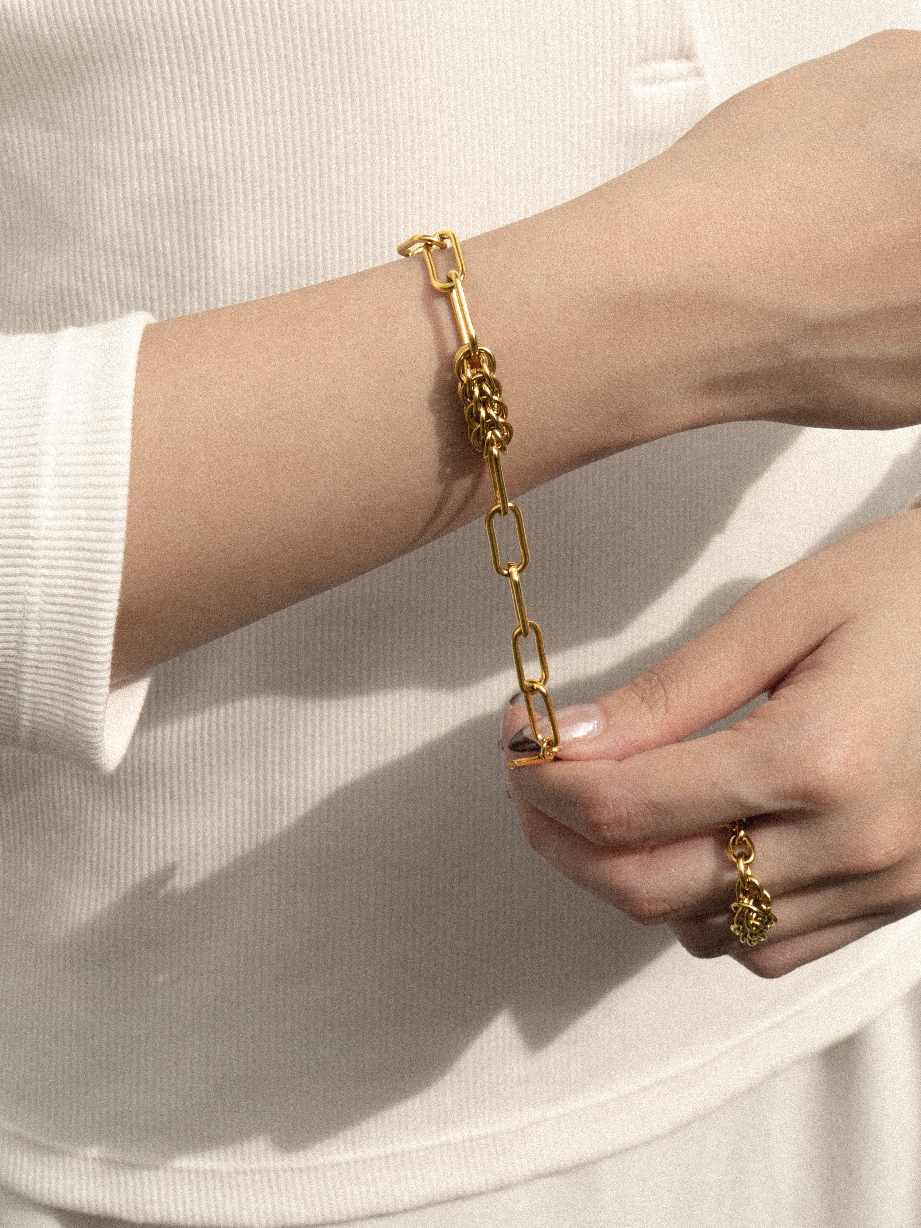 A close-up of a person's wrist and hand wearing a gold chain bracelet with a textured central link, paired with a matching gold ring on the finger.
