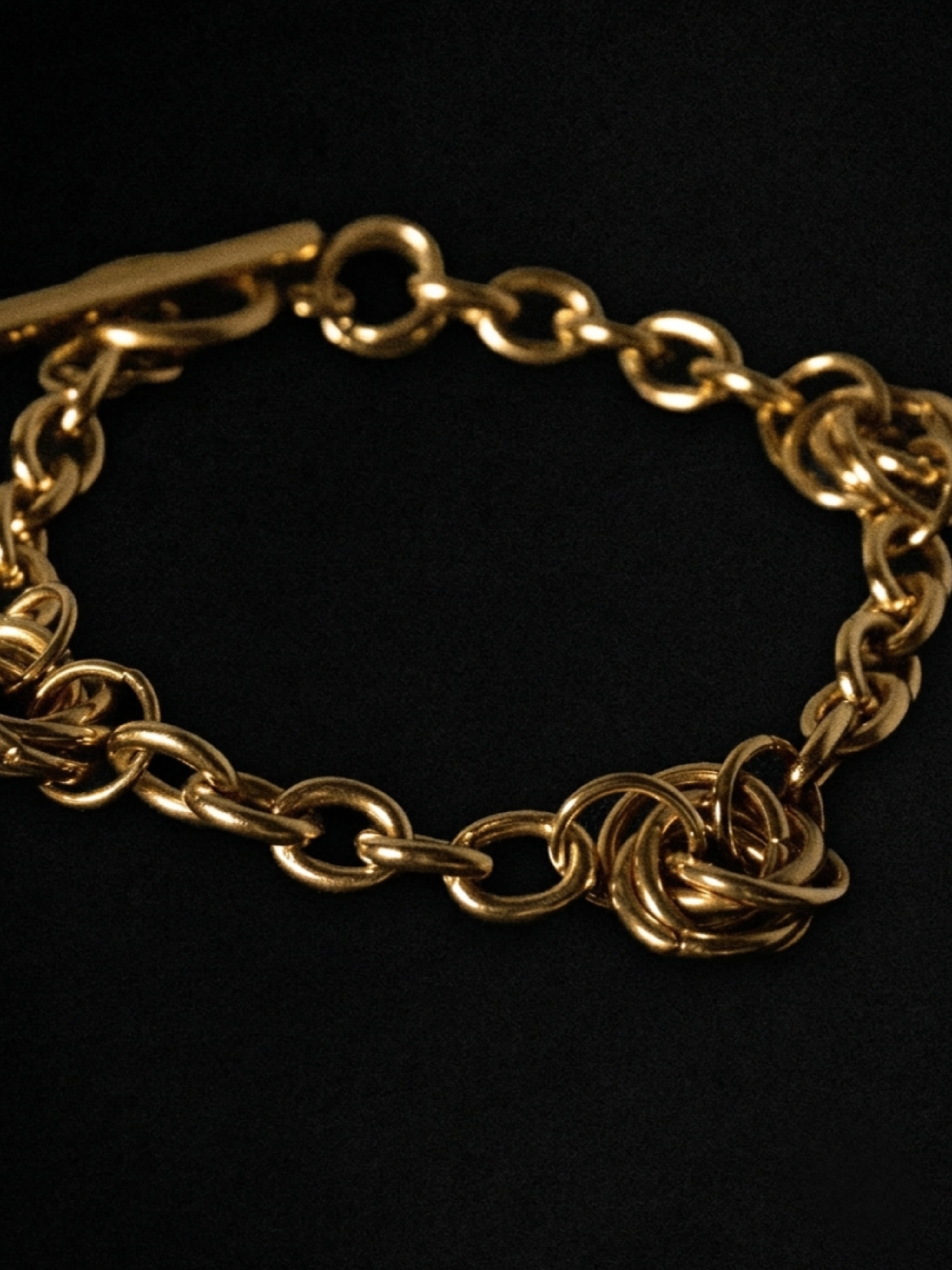 A close-up of a gold chain bracelet with interlocking links and a clasp, photographed against a solid black background.