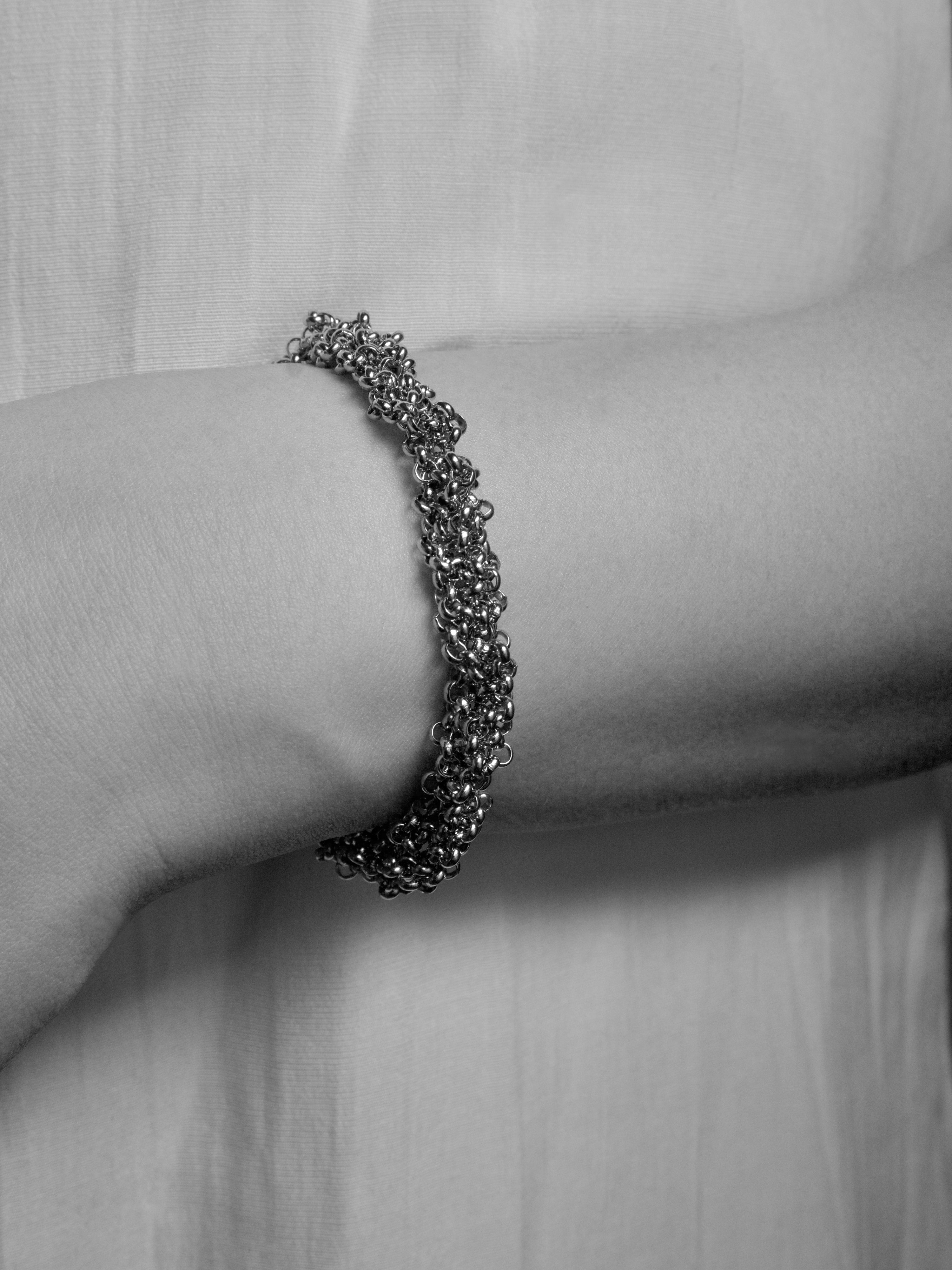 A close-up of a textured metallic chain bracelet worn on a person's wrist, shown in a black and white photograph against a soft fabric background.