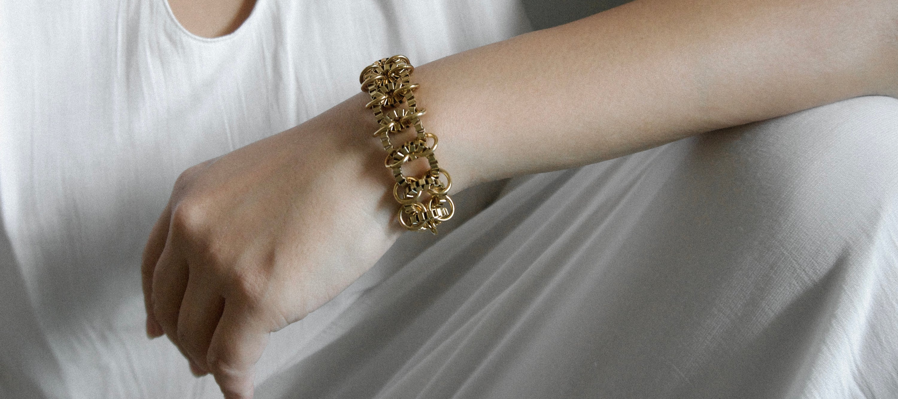 A close-up of a person's wrist wearing a chunky, textured gold chain bracelet, dressed in a white top and white draped fabric.