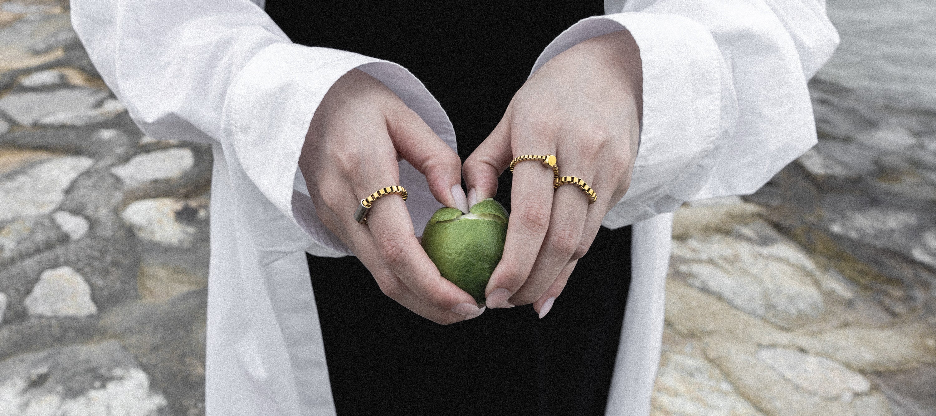 A close-up of a person's hands holding a green apple, wearing a white shirt over a black dress and adorned with gold rings, set against a blurred stone background.