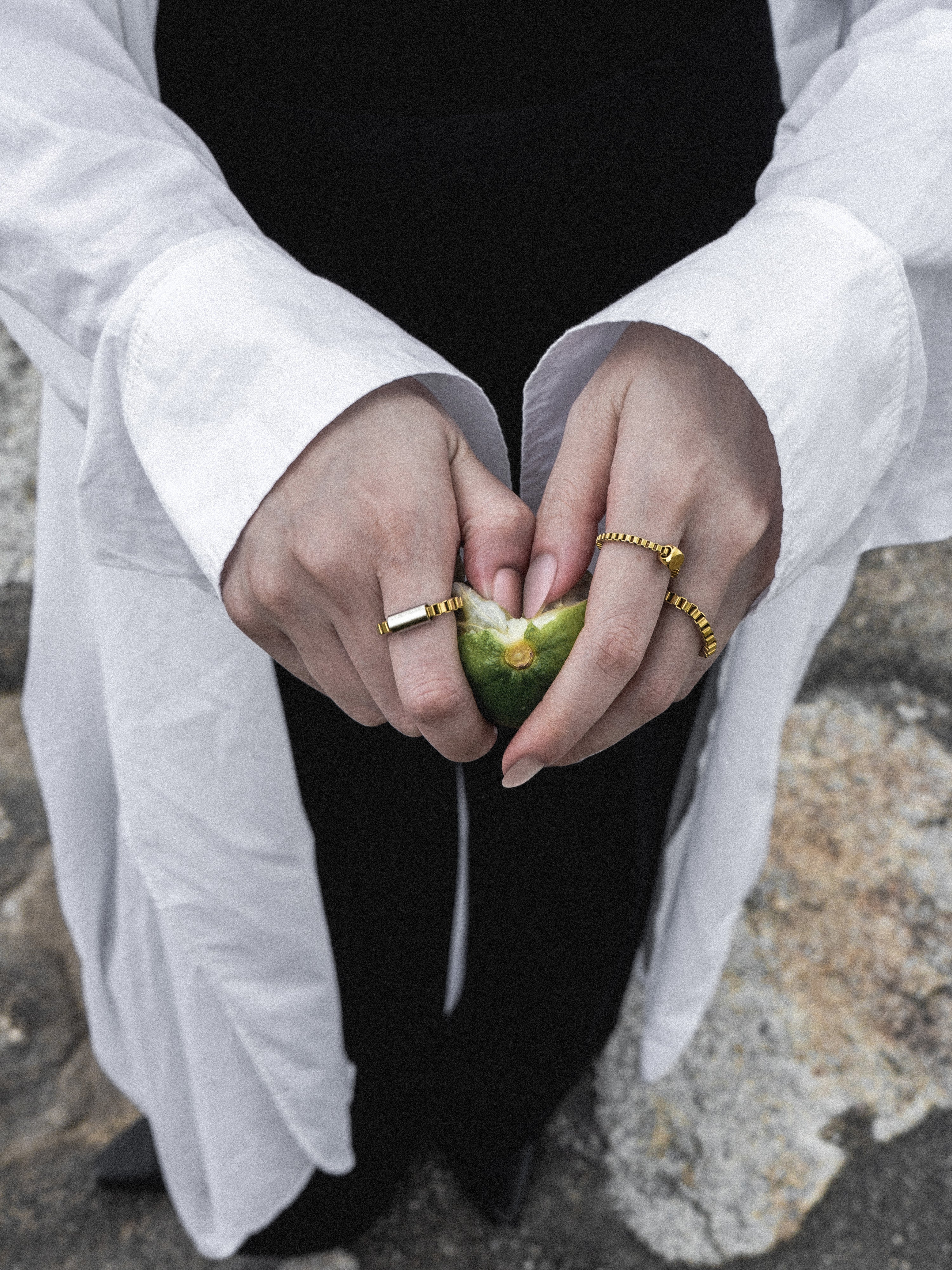 A close-up of a person's hands wearing several gold rings, including a wide gold band with a rectangular stone and a textured gold ring, while crushing a green lime. The person is wearing a white shirt and black skirt against a rocky outdoor background.