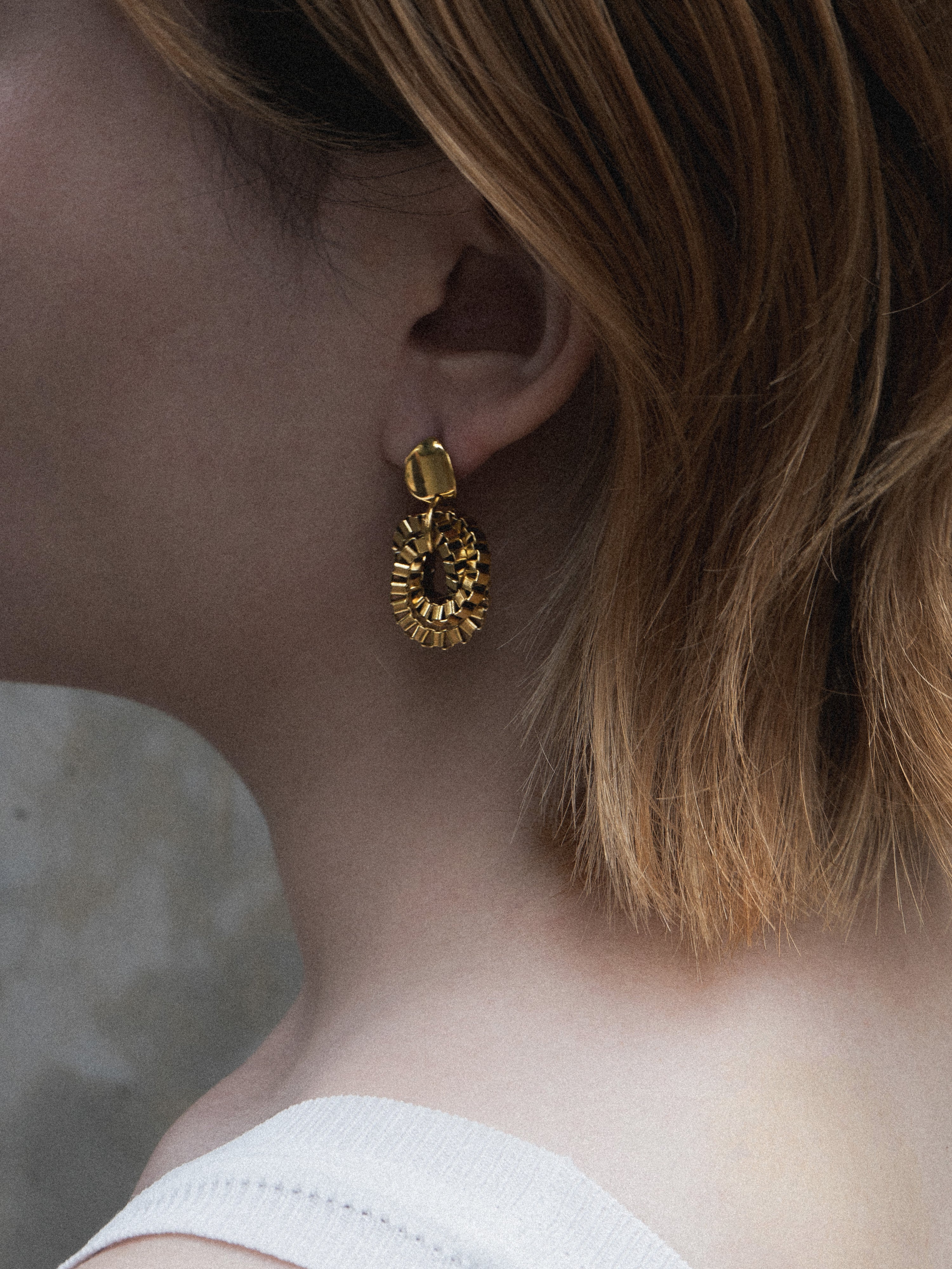 Close-up of a person wearing a gold earring with chains interlocking detail against a concrete wall 