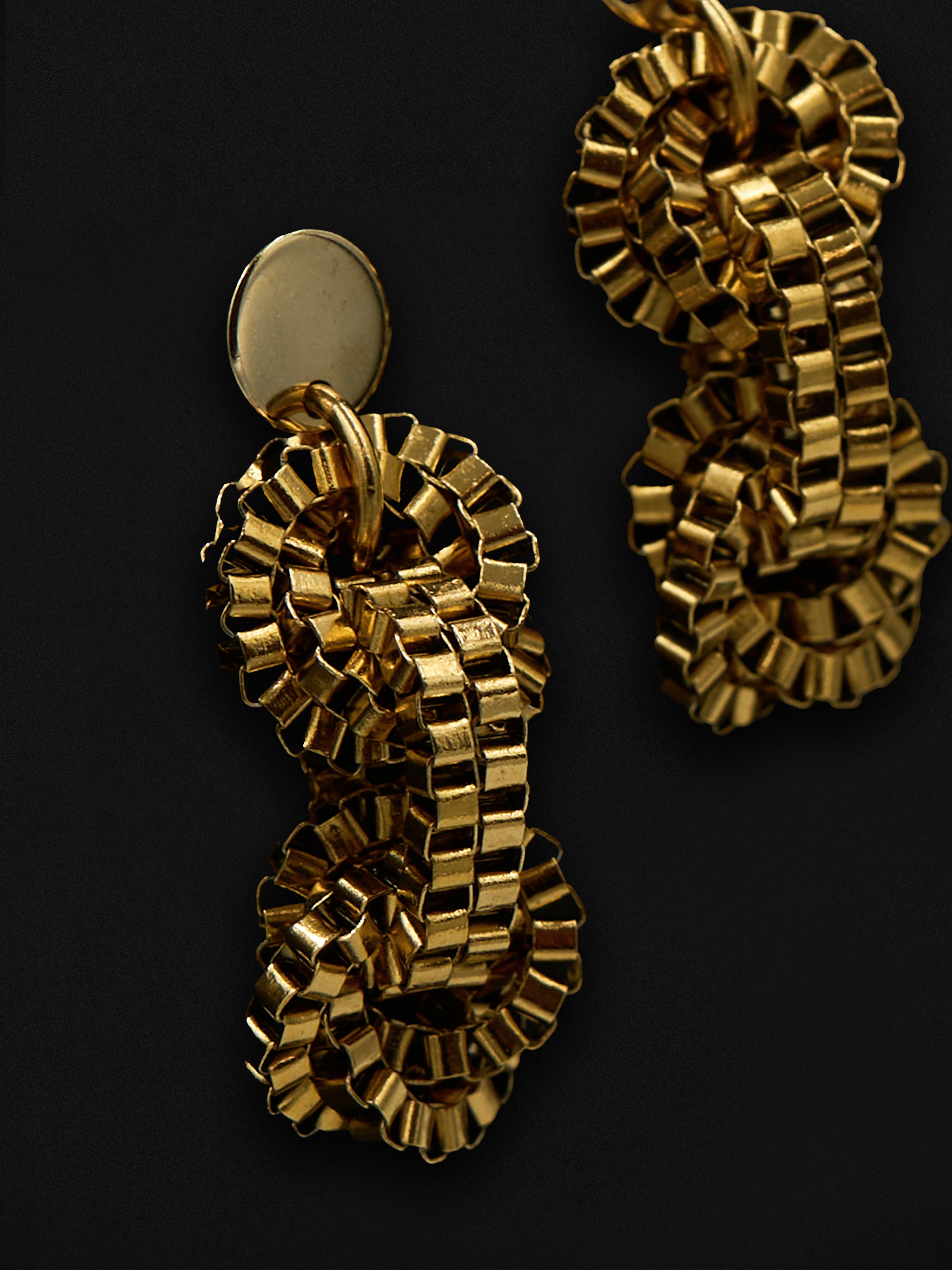 A pair of chunky gold-tone statement earrings with a textured, chain-like link design and a flat oval disc at the top, shown against a solid black background.