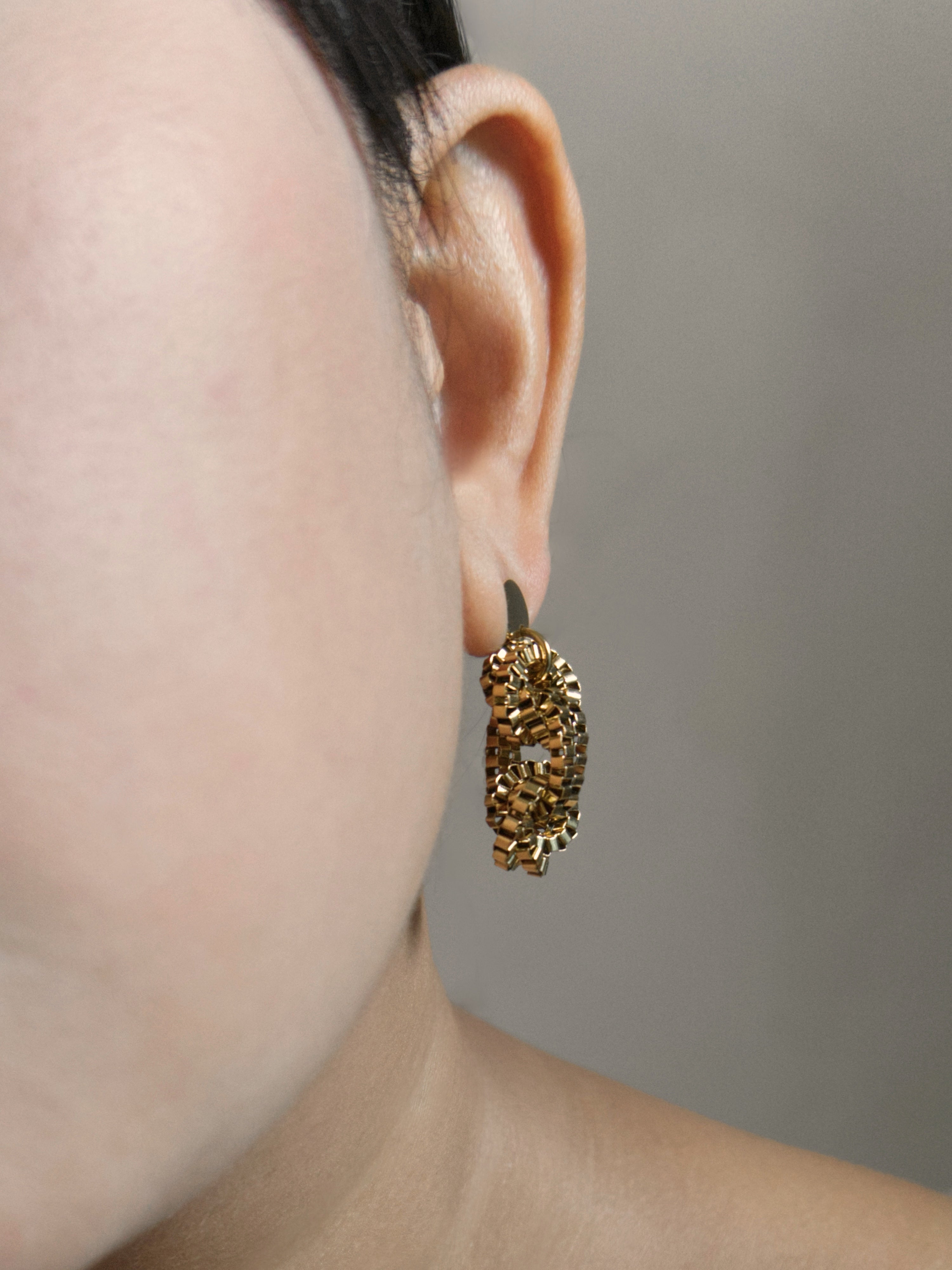 A close-up lifestyle shot of a person from behind, showing them wearing textured gold dangle earrings and a white t-shirt against a textured beige wall.