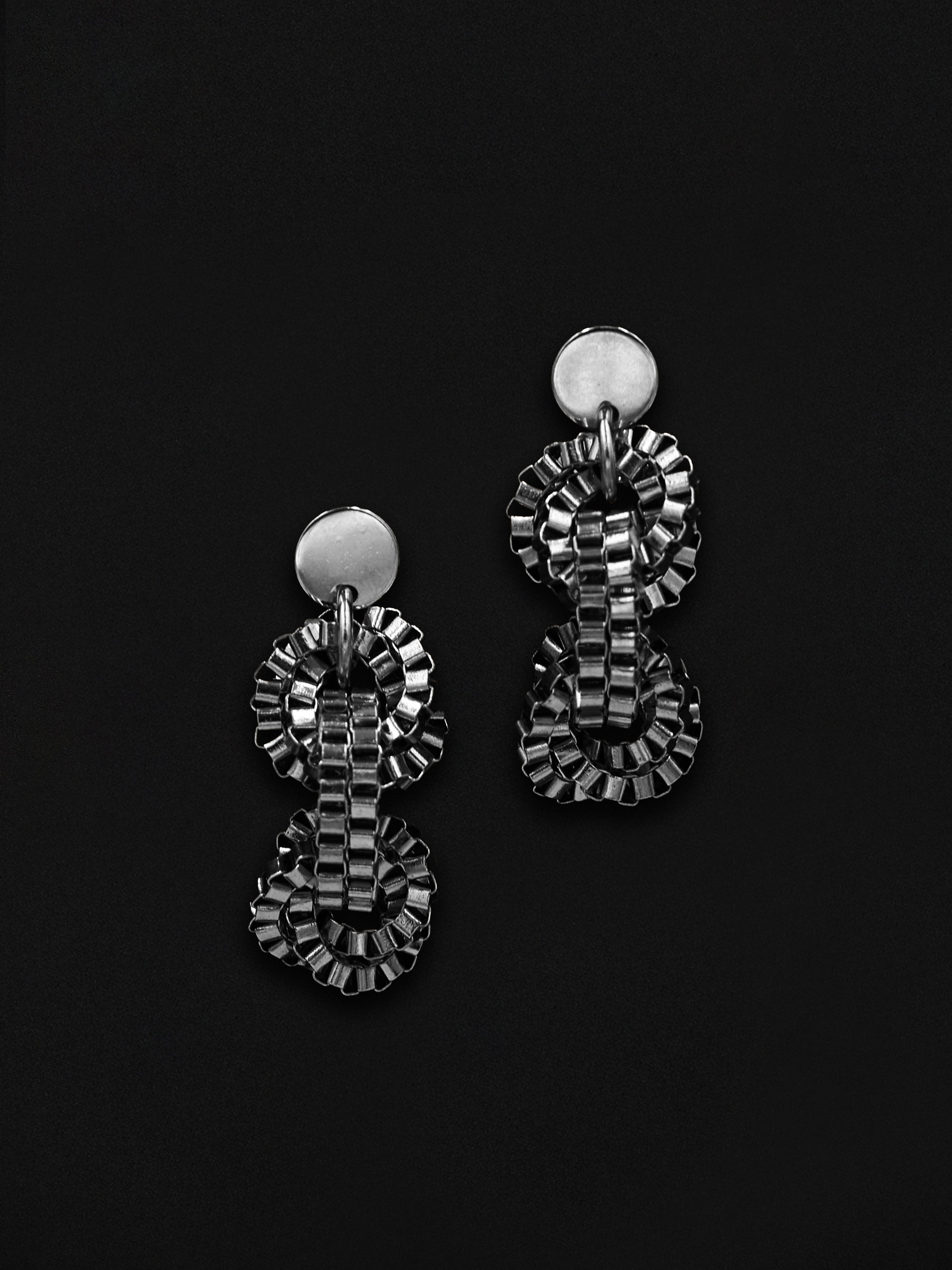 A pair of silver-toned statement dangle earrings with textured, layered geometric loops and round stud backs, displayed on a solid black background.