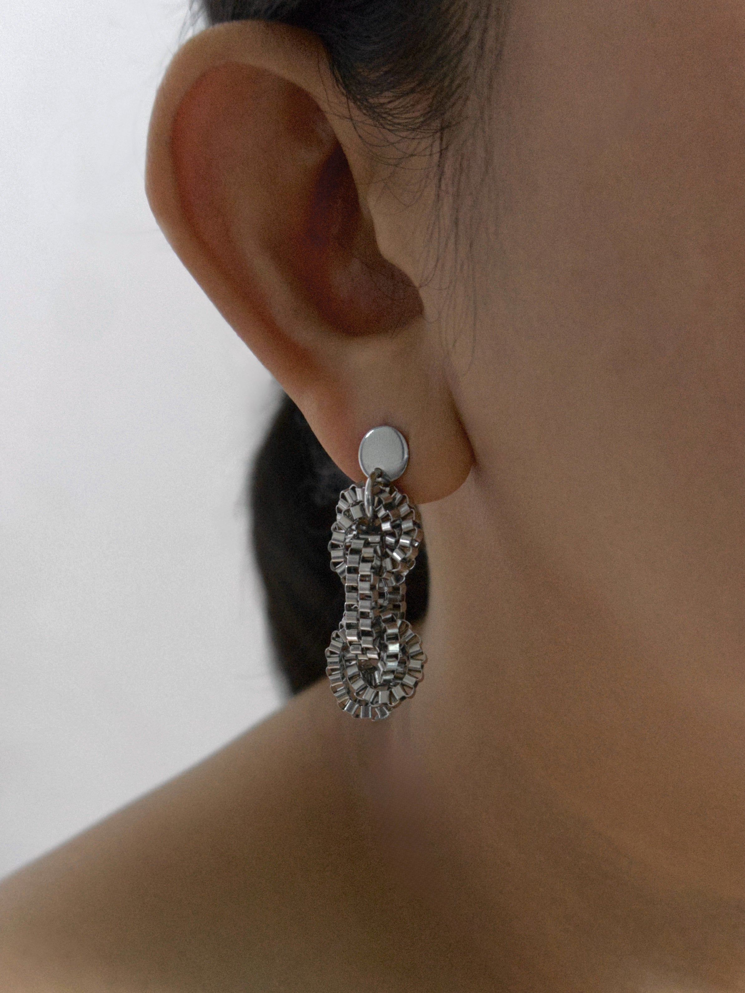 A model with hair tied wearing silver interlocking chain earrings on one side against a light neutral background.