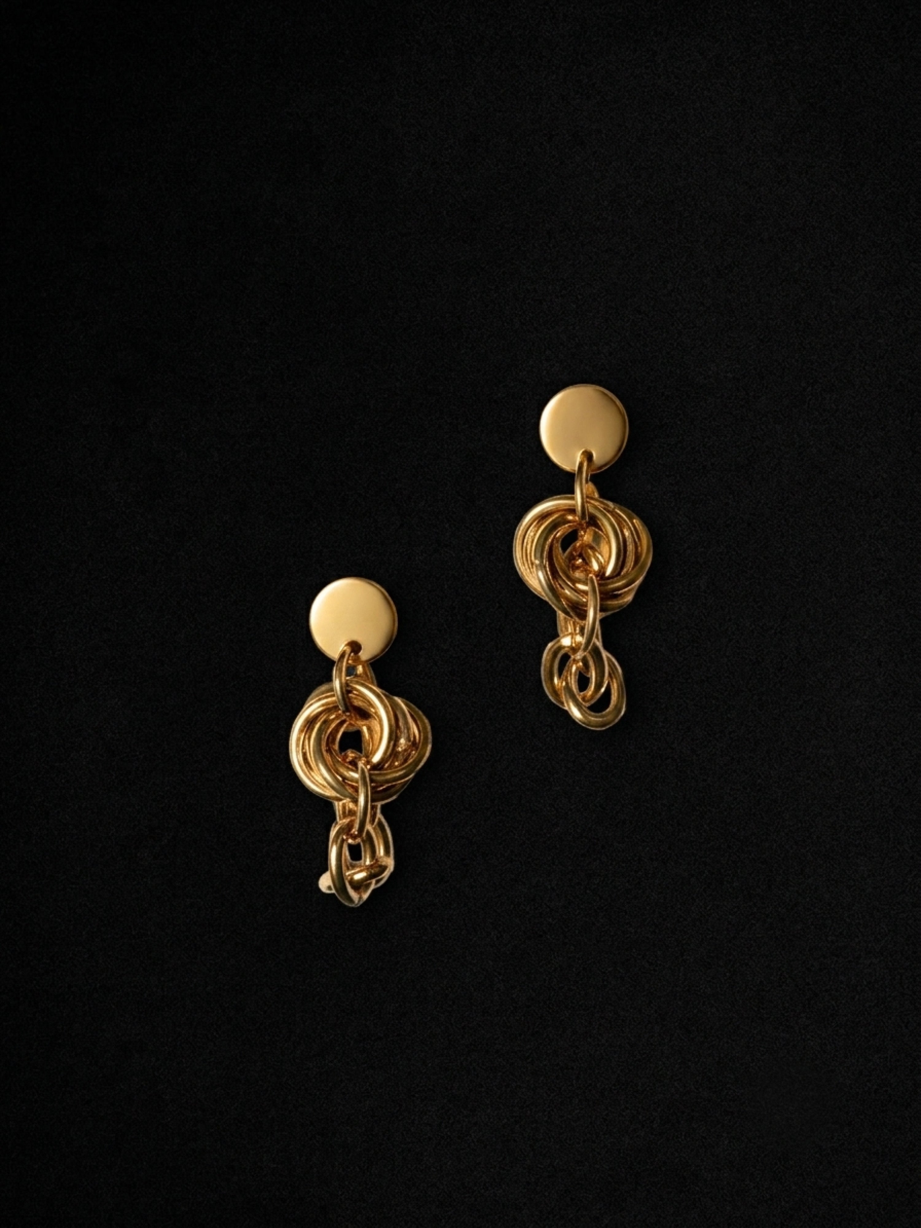 A pair of gold-toned statement earrings with circular studs and layered, chain-like dangling links, displayed on a solid black background.