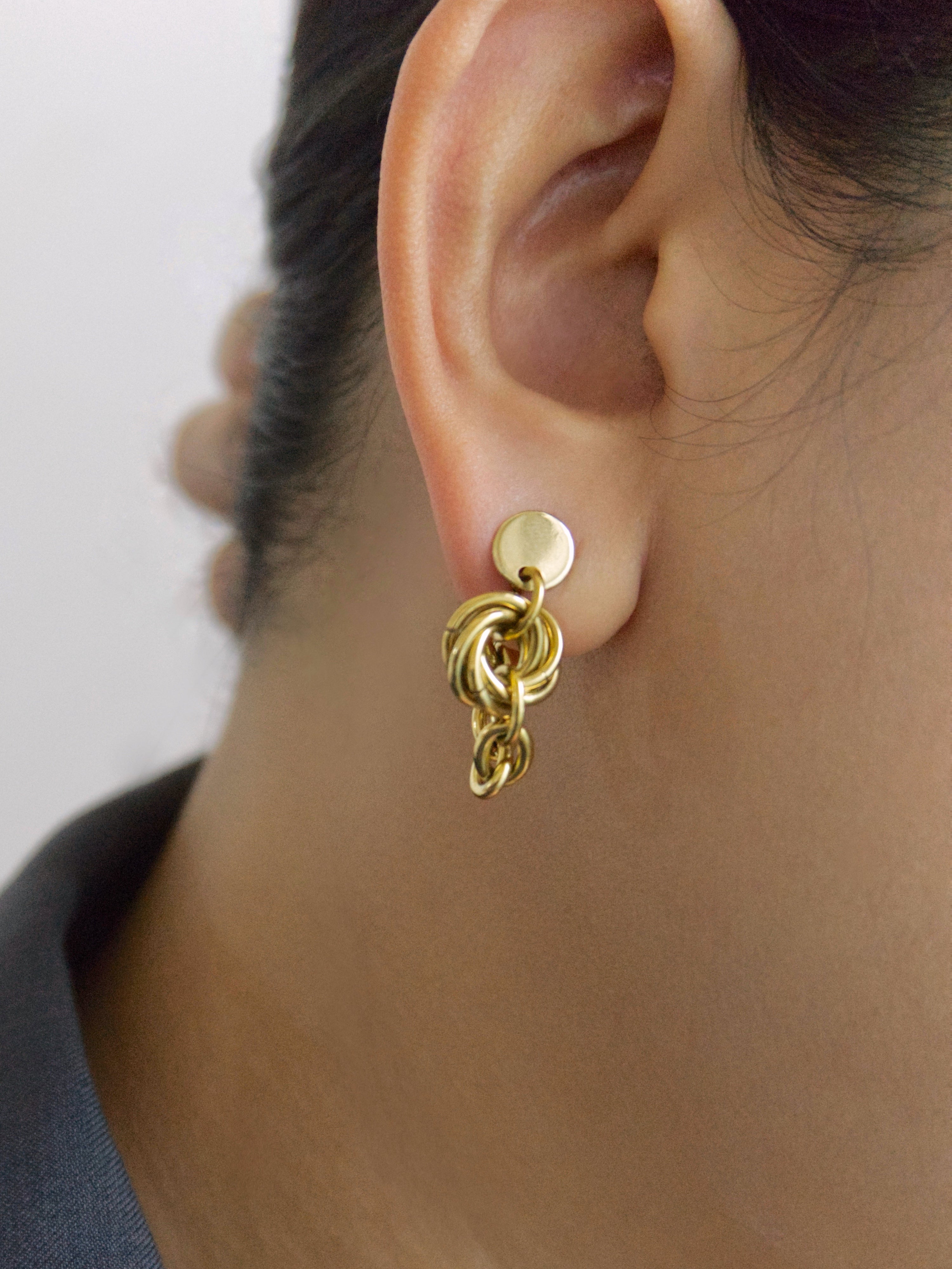A close-up of a person's ear wearing a small gold hoop earring with a dangling interlocking circular charm, against a light gray background.