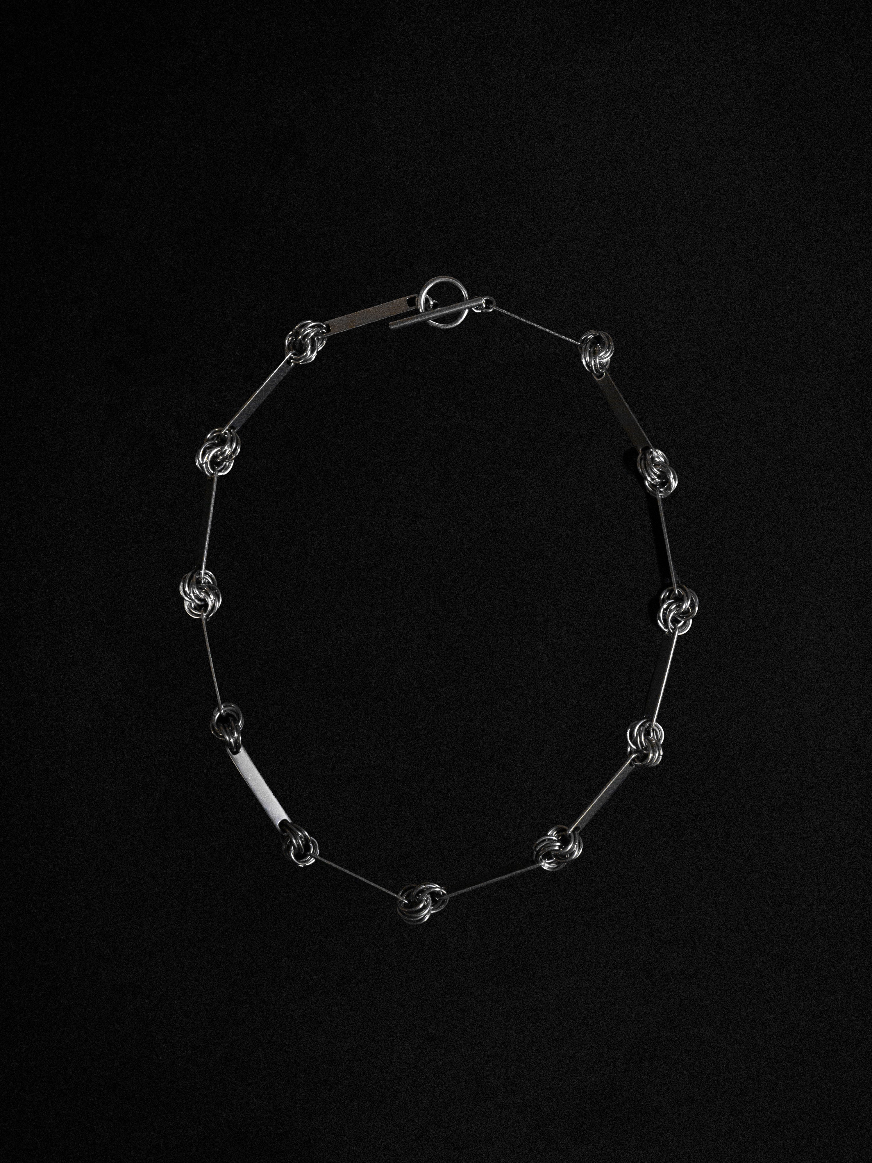 A silver-colored necklace with a chainmaille pattern and rectangular hardware segments, displayed against a black background.