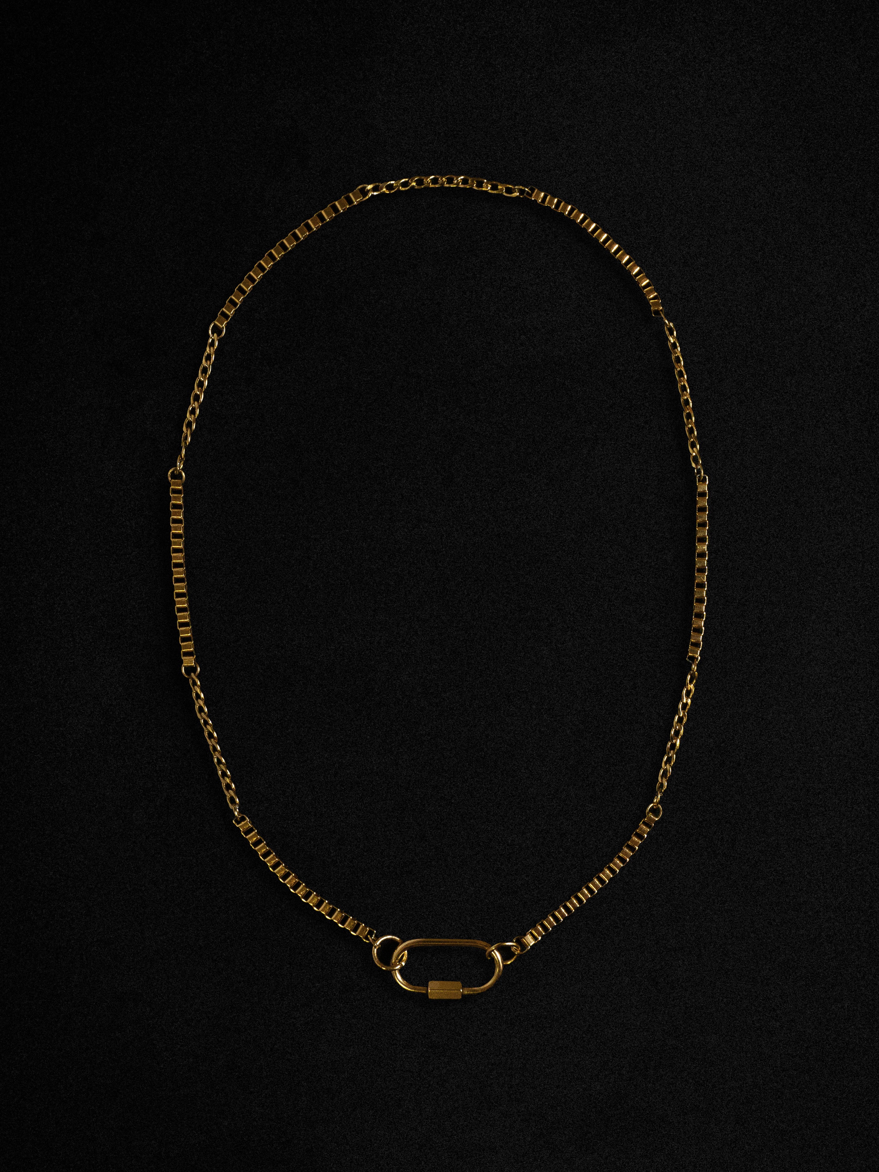 A gold-colored necklace with a carabiner clasp, laid out on a black background.