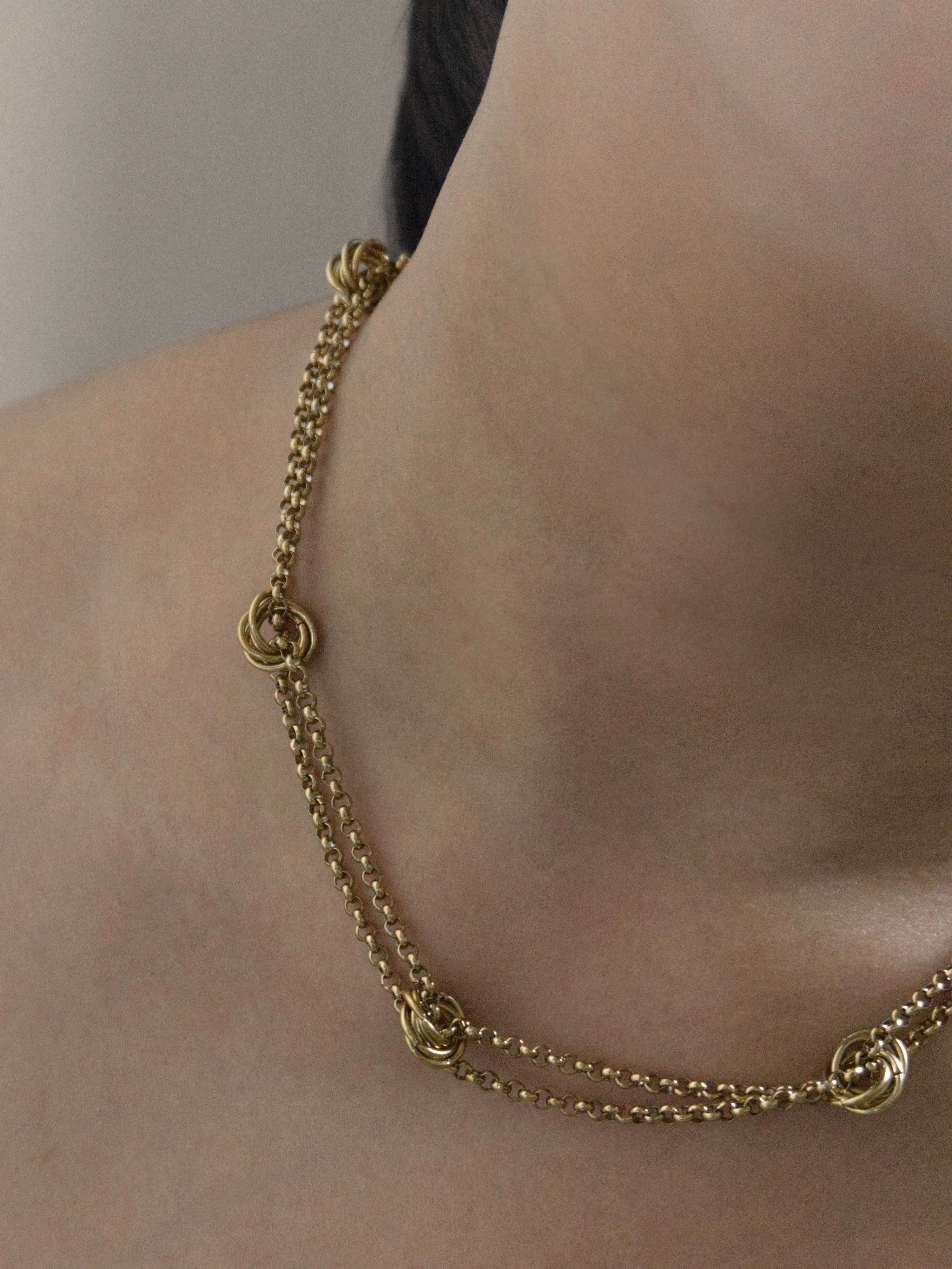 A person wearing a gold necklace with twisted rings at intersection against a neutral background.