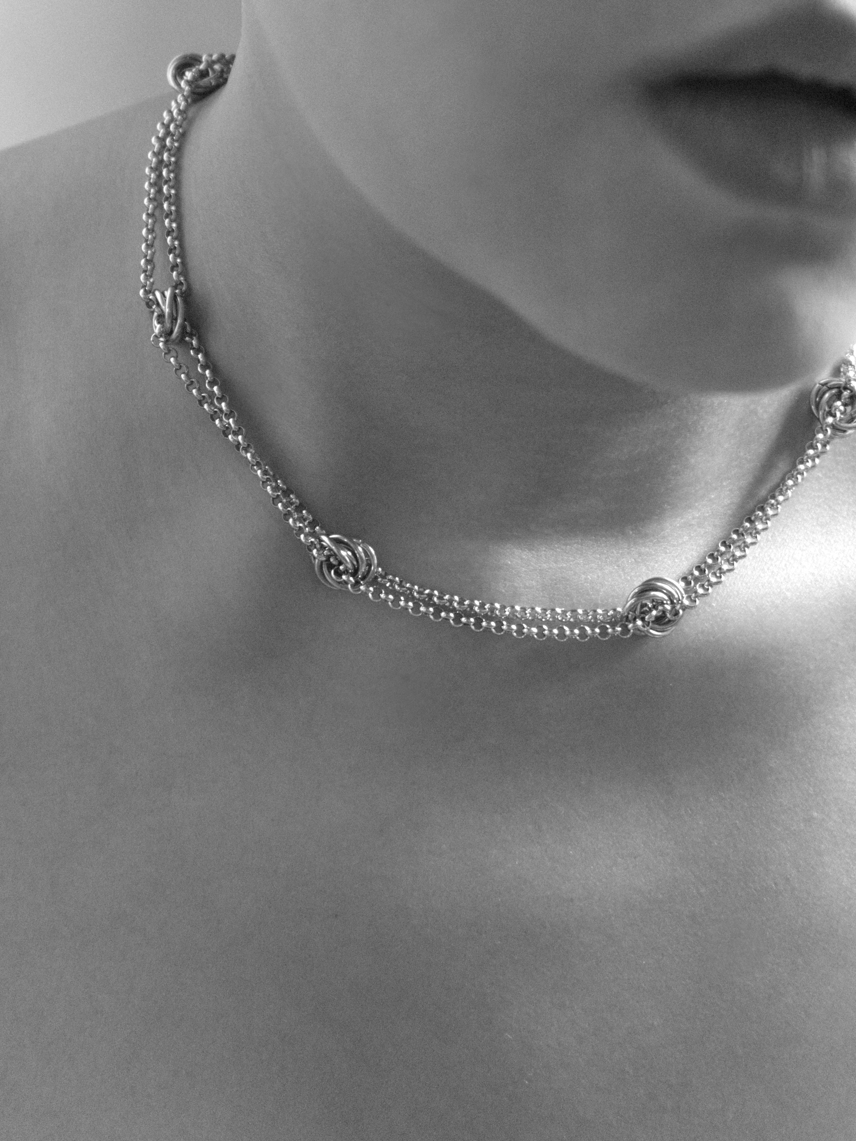 A close-up black and white photograph of a model's neck and lower face wearing a delicate chain short necklace with interlocking circular links.