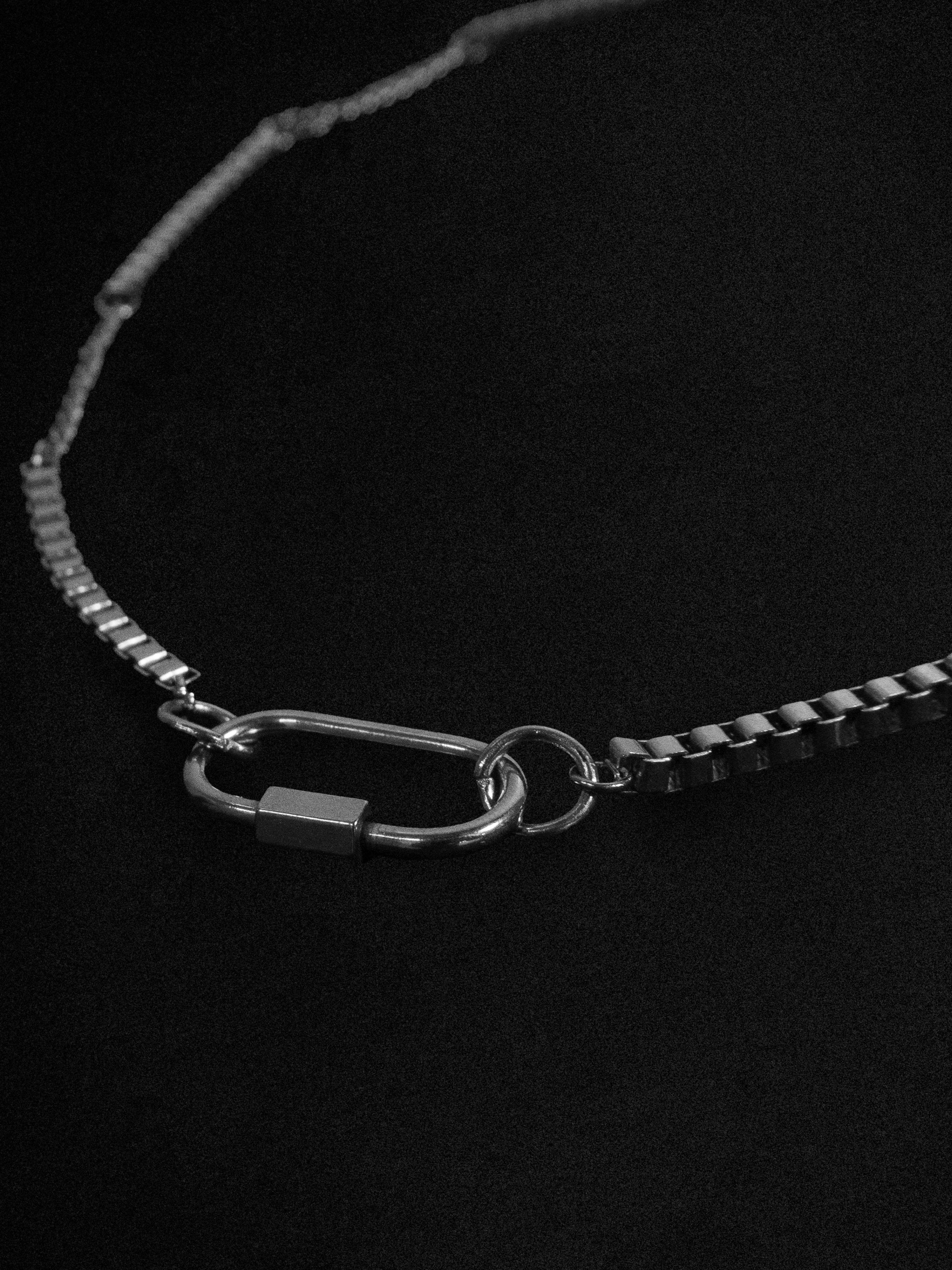 A silver-toned metal chain necklace with a distinctive carabiner-style clasp and a small rectangular lock, displayed on a dark black background.