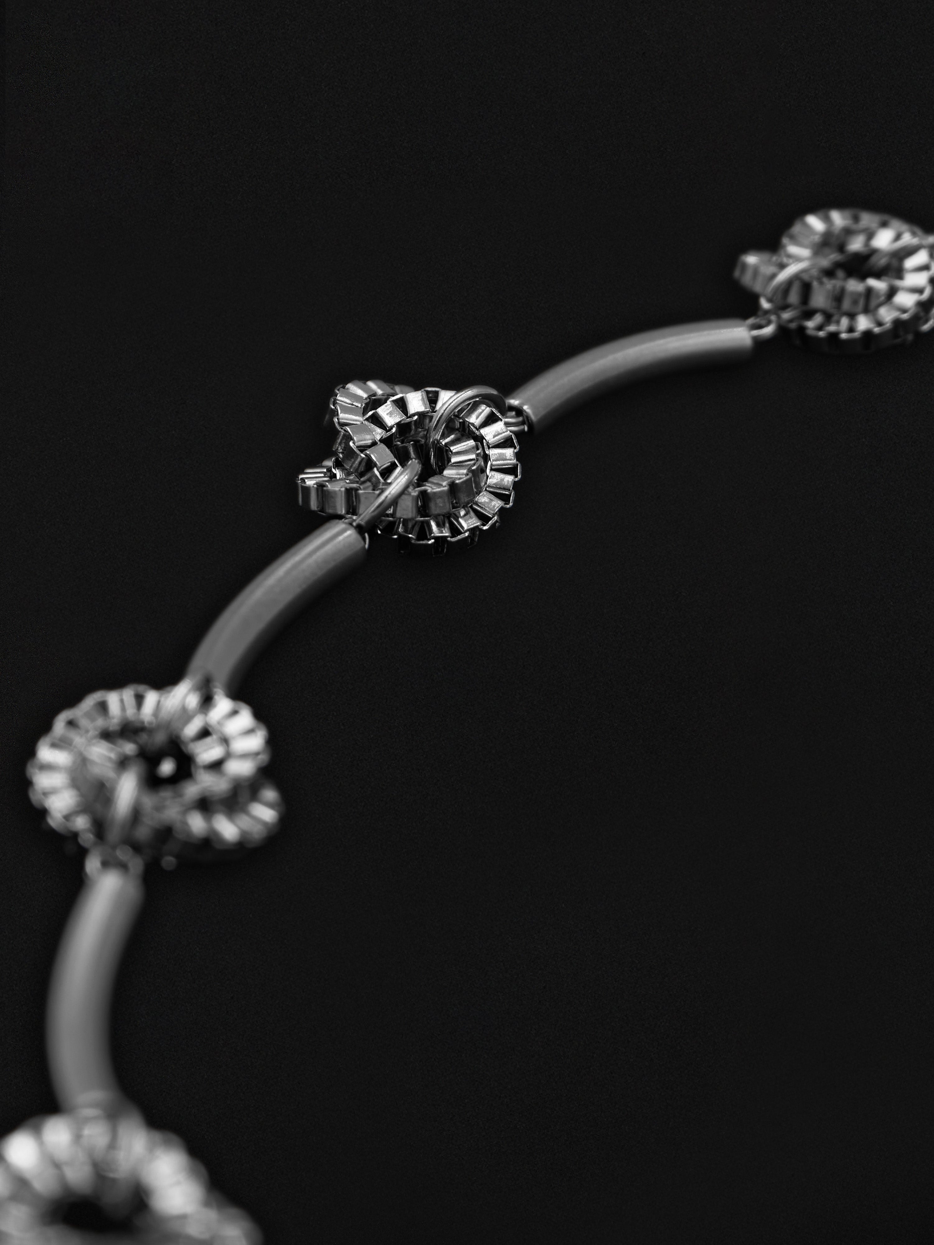 A close-up of a silver-toned curved chain necklace with ornate flower-like links, photographed against a solid black background.