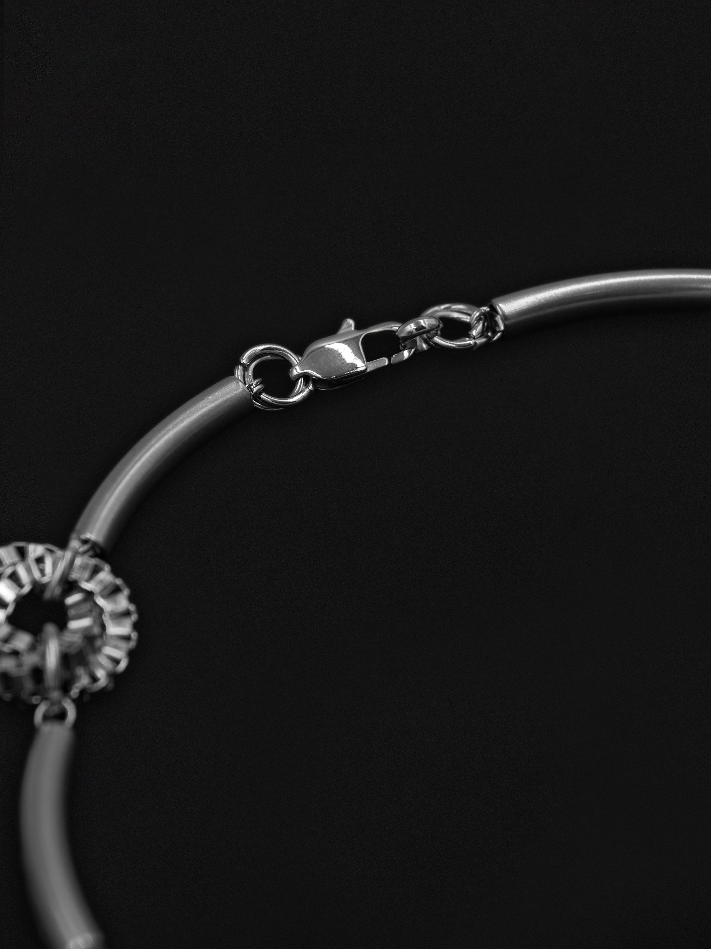 A close-up of a silver-toned metal necklace showing a lobster clasp, chain links, and part of a decorative charm against a solid black background.