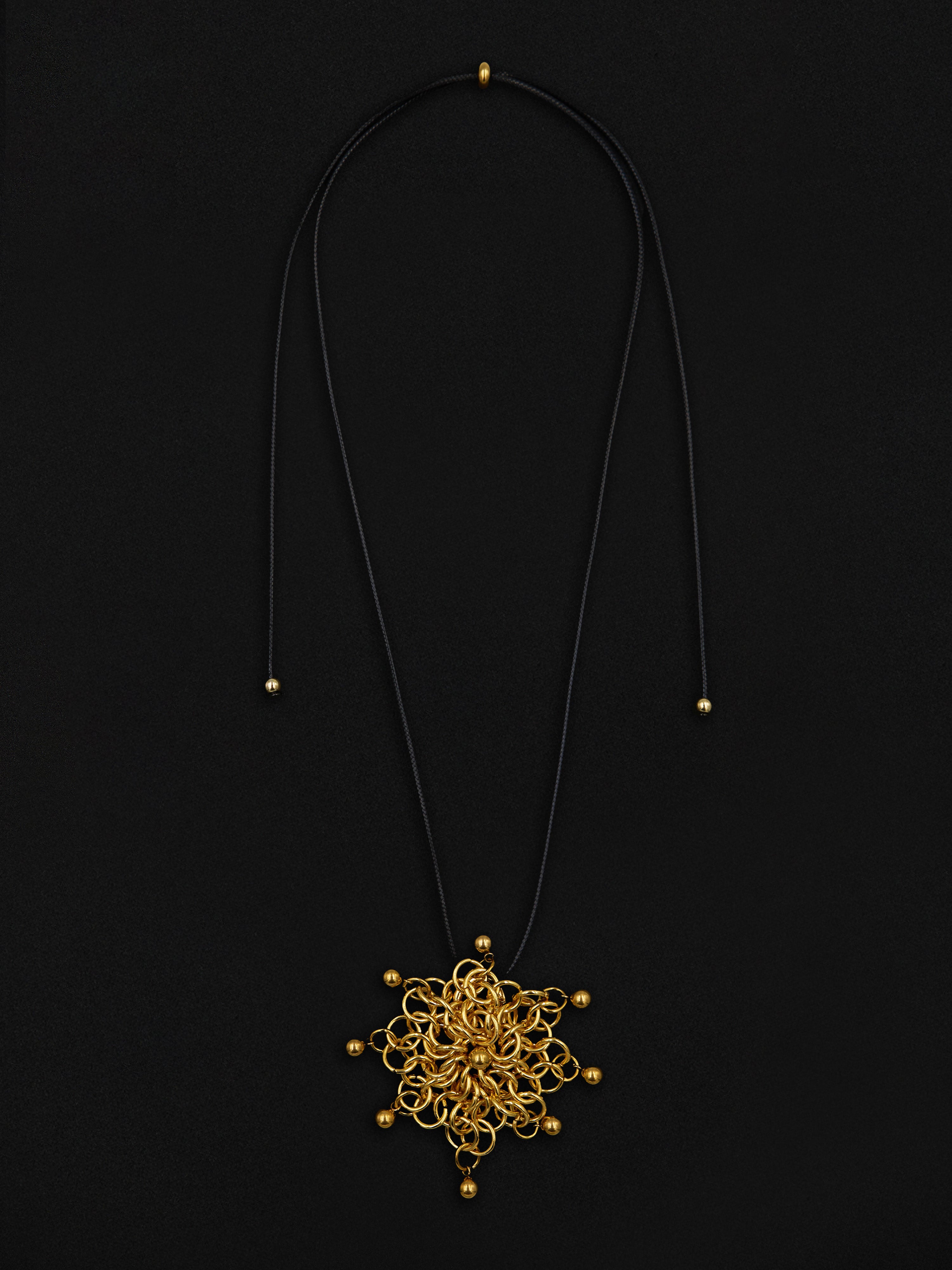 A black cord necklace featuring an intricate, circular gold filigree pendant with small gold beads, displayed against a solid black background.