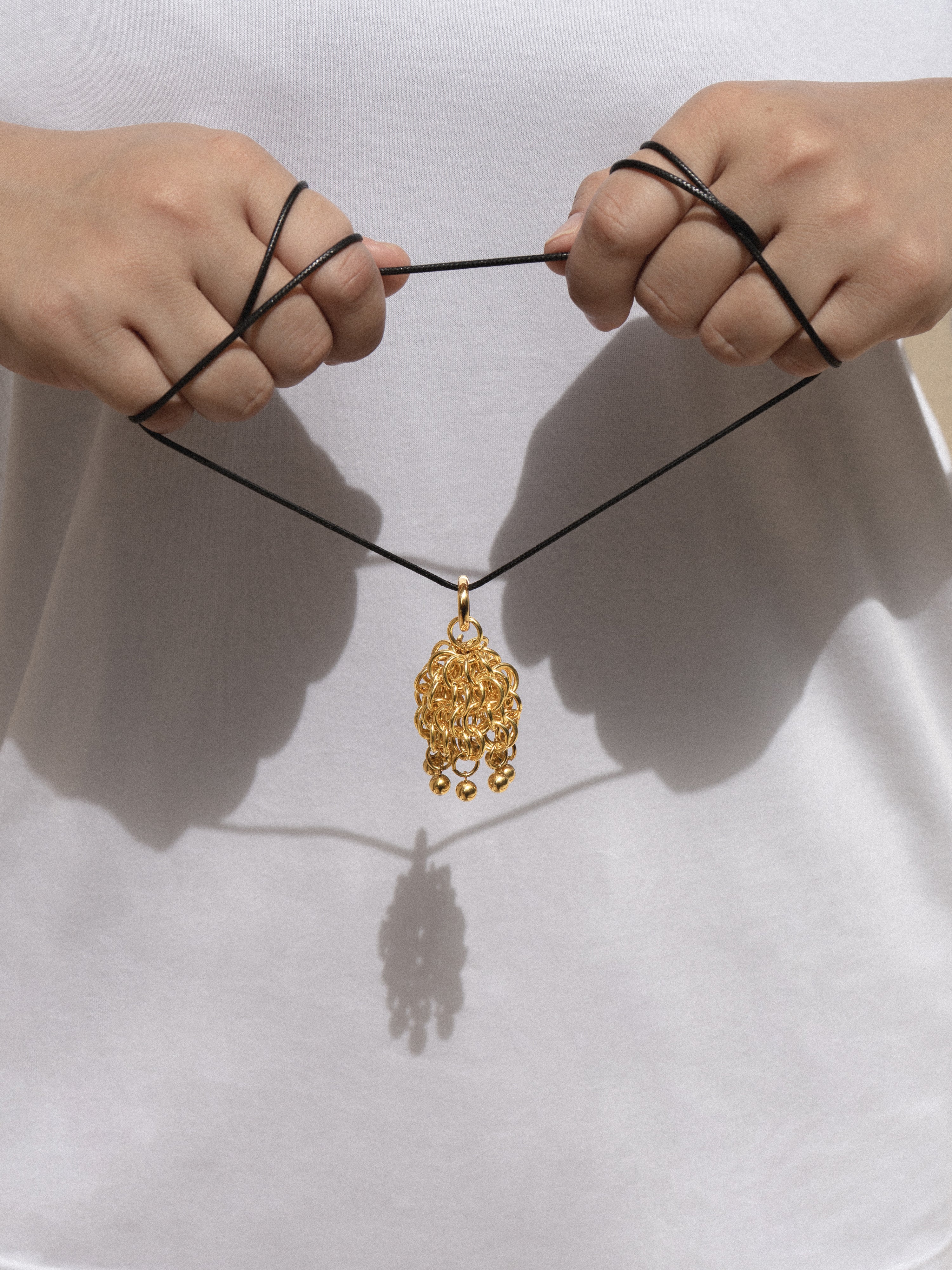 A person's hands holding a black cord necklace with a detailed gold pendant against a white shirt background.