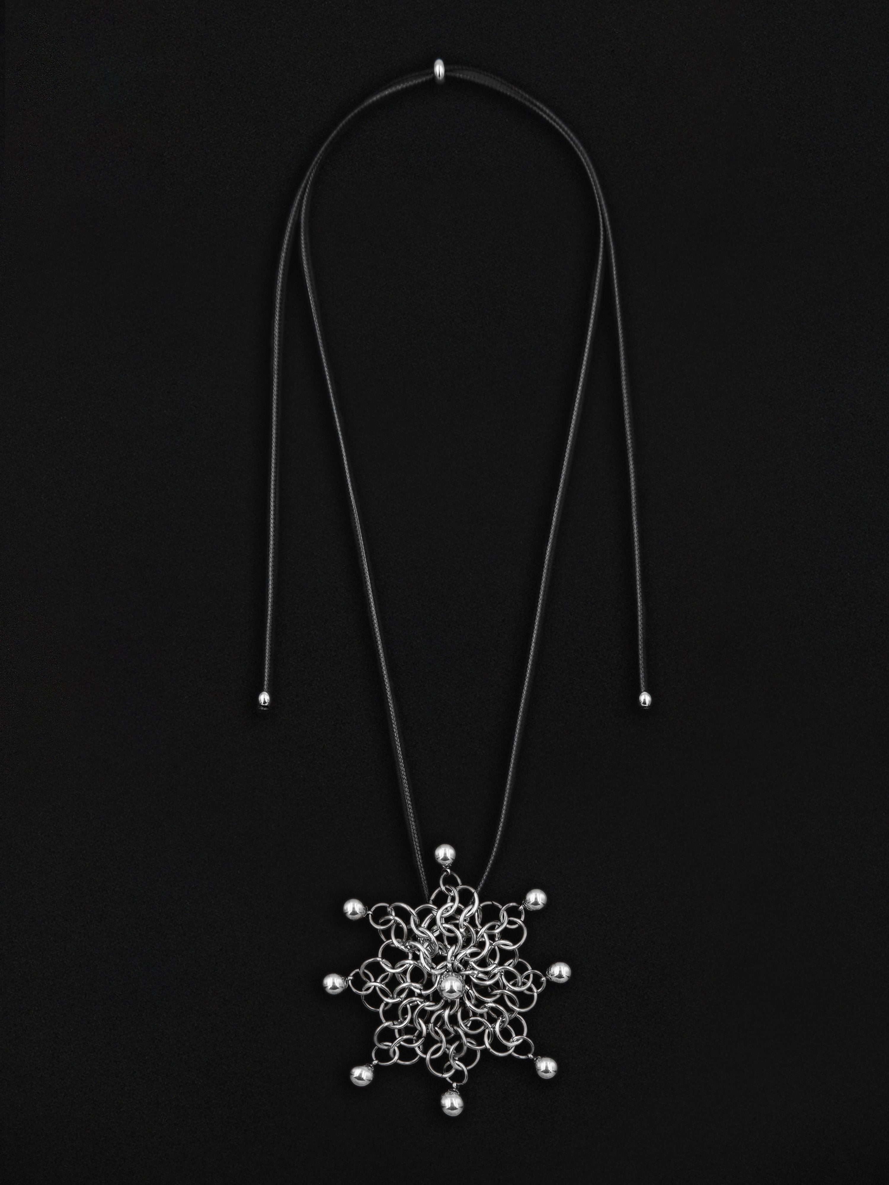 A long black cord necklace with a silver-toned filigree pendant and small bead accents, displayed against a solid black background.