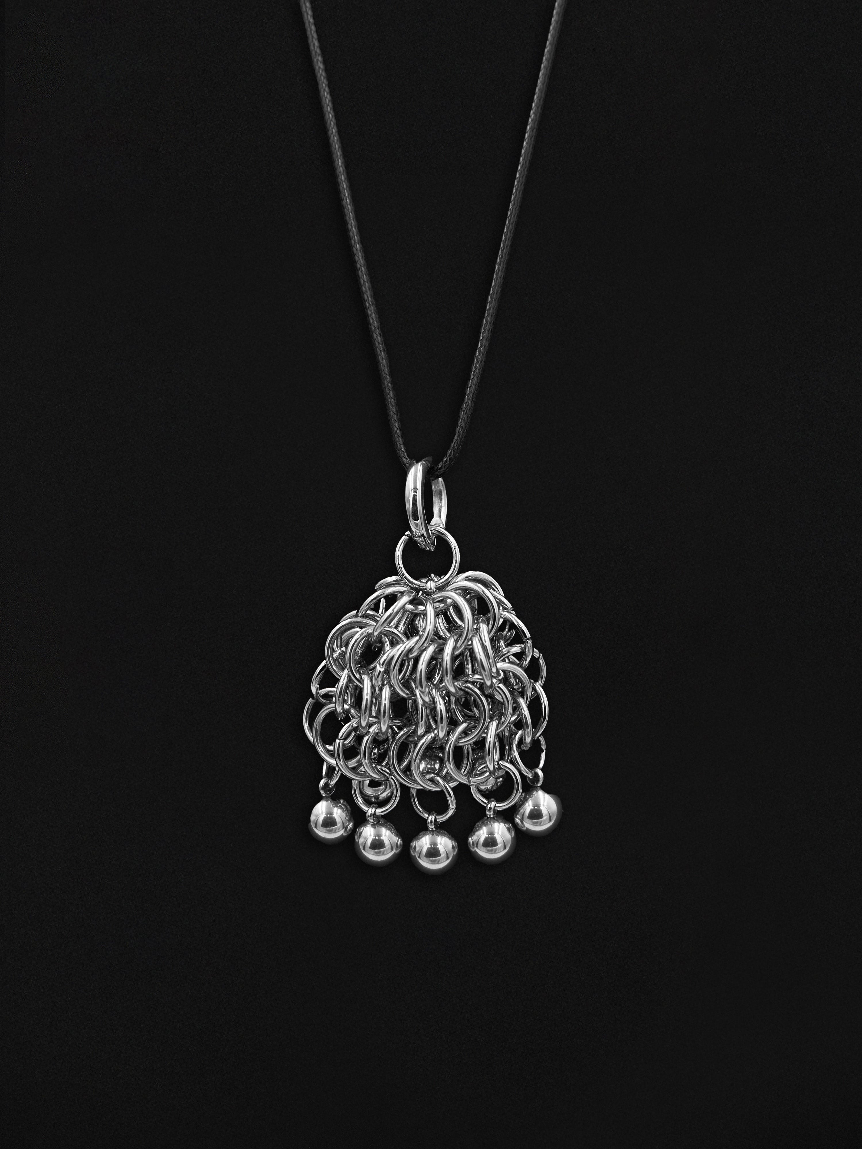 A silver-toned pendant necklace featuring a dense, textured mesh-like cluster of interlocking rings with five small dangling bell charms, suspended from a thin black cord against a solid black background.