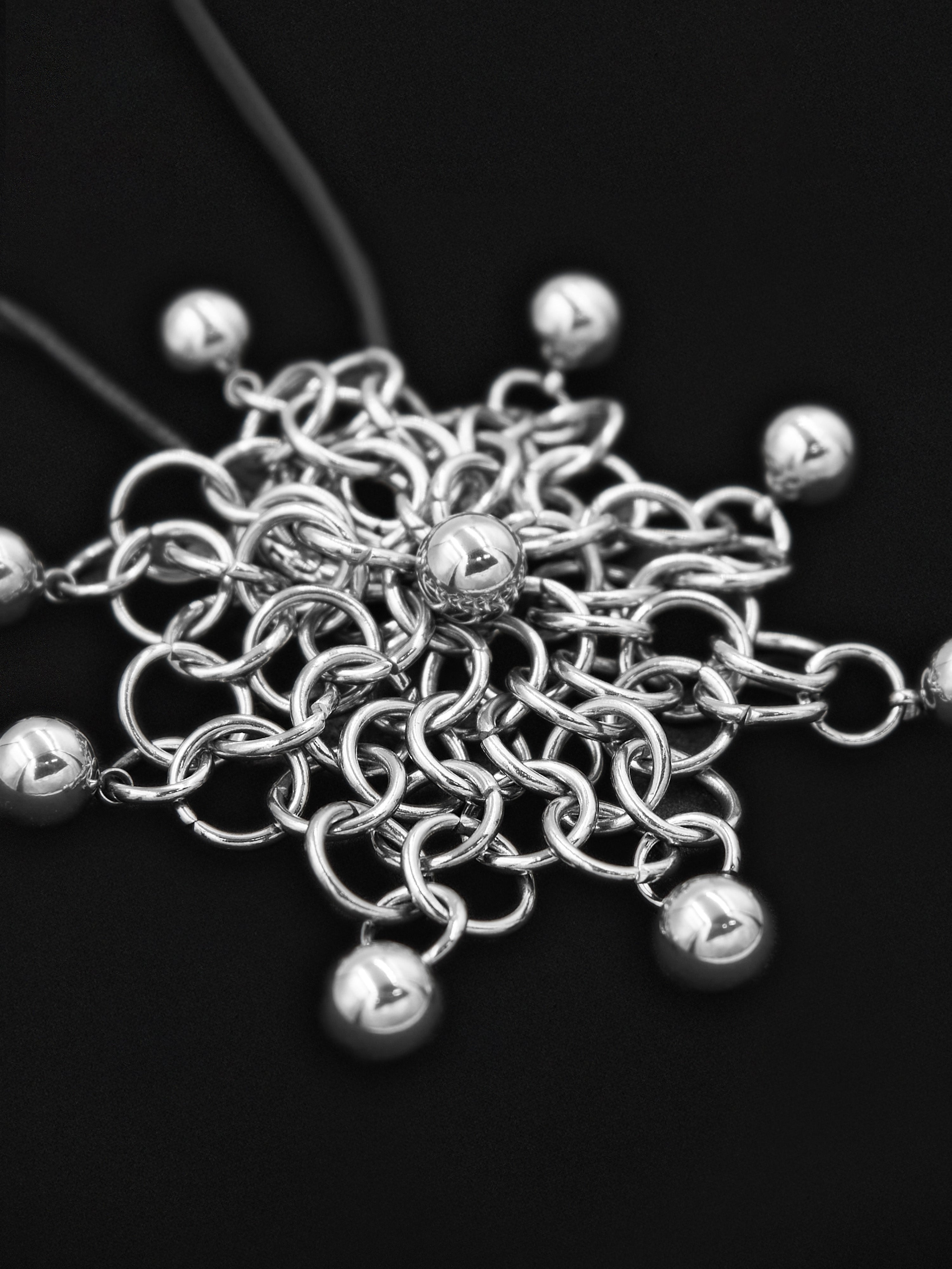 A close-up of a silver-toned chainmail-style pendant or charm with multiple dangling polished beads, photographed on a black background.