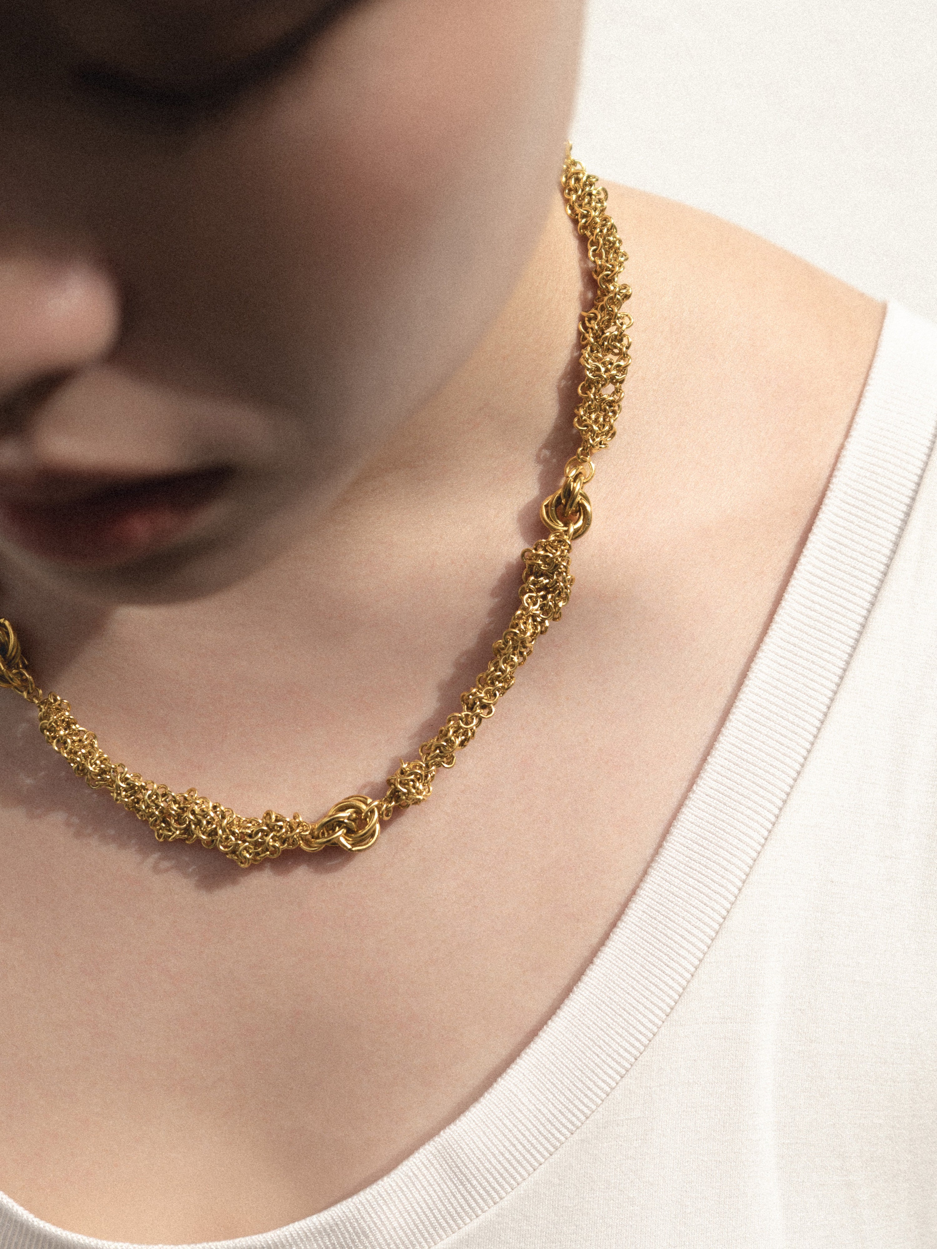 A close-up of a person wearing a textured gold chain necklace with a distinctive interlocking knot design, paired with a white ribbed top.